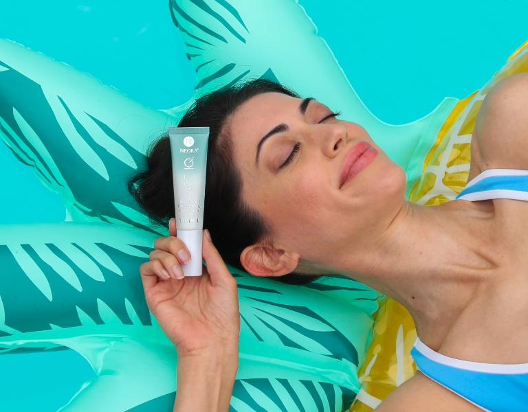 Woman laying on a pineapple floaty in the pool and holding Neora’s Invisi-Bloc SPF40 Sunscreen Gel.